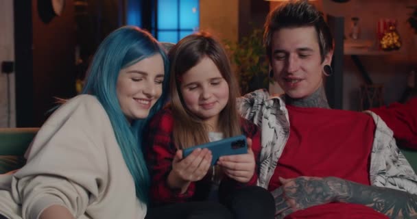 Happy family watching funny cartoon and laughing while sitting on sofa .Modern mom, dad and their daughter looking at smartphone screen in horozontal landscape mode and smiling at home . — стоковое видео