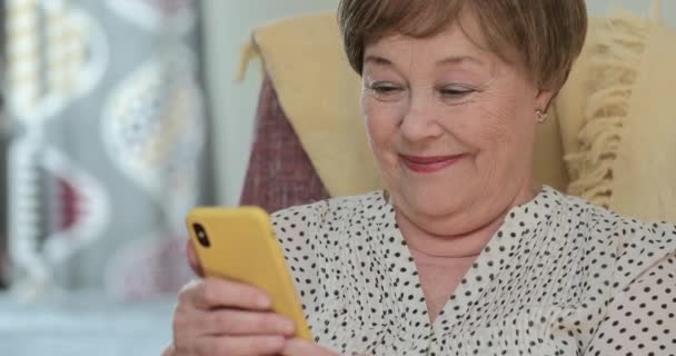 Mujer hermosa madura mirando la pantalla del teléfono inteligente y sonriendo mientras recibe un mensaje divertido. Vieja usando su teléfono móvil moderno y recibiendo buenas noticias mientras está sentada en casa . — Vídeo de stock