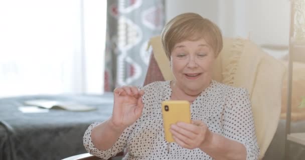 Ältere schöne Frau in den 60ern, die auf den Smartphone-Bildschirm schaut und lächelt, während sie zu Hause sitzt. Alte moderne Dame benutzt ihr Handy und bekommt gute Nachrichten, während sie ihre Freizeit verbringt. — Stockvideo