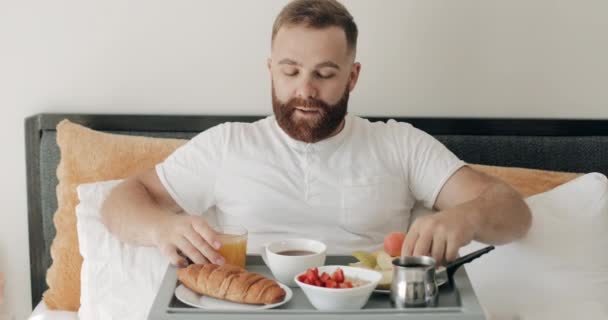 Jovem homem sorrindo sentado com bandeja cheia de comida em suas pernas e desfrutando de manhã. Bonitão comendo maçã e segurando um copo de suco enquanto toma café da manhã na cama. Fundo de casa . — Vídeo de Stock