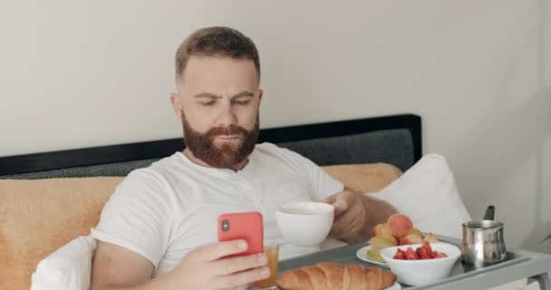 Hombre barbudo guapo usando su teléfono inteligente mientras bebe café por la mañana. Chico de 30 años mirando la pantalla del teléfono mientras desayuna y se sienta con la bandeja llena de comida en la cama . — Vídeos de Stock