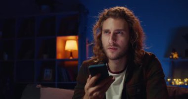 Genç yakışıklı sakallı adam kanepeye otururken sanal asistan uygulaması kullanıyor. Bin yıllık uzun saçlı adam akıllı telefonunu kullanırken ekranda konuşuyor ve dokunuyor. Ev arkaplanı.