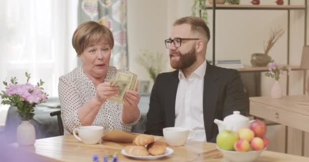 Chockad mogen kvinnlig pensionär räknar sedlar och ser tacksamt på sin vuxne son. Ung affärsman stödja sin pensionerade mor med en stor summa pengar när du besöker henne. — Stockvideo