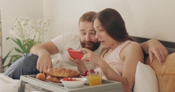 Man and woman making big eyes while looking shocked about unbelievable news on smartphone. Young couple with amazed face expression sitting on bed during breakfast. — Stock Video