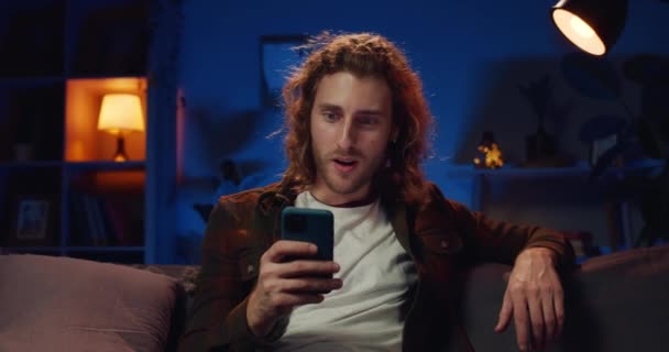 Millennial baardman zeggen wow en het maken van ja gebaar, terwijl het kijken naar smartphone scherm. Goed uitziende langharige man die wint terwijl hij op de bank zit. Concept van succes en goed nieuws. — Stockvideo