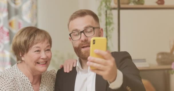 Veselá důchodkyně a dospělý vnuk v brýlích pro selfie a dívají se na obrazovku telefonu. Usmívající se syn fotografování s matkou na přední kameru smartphone a usmívání. — Stock video
