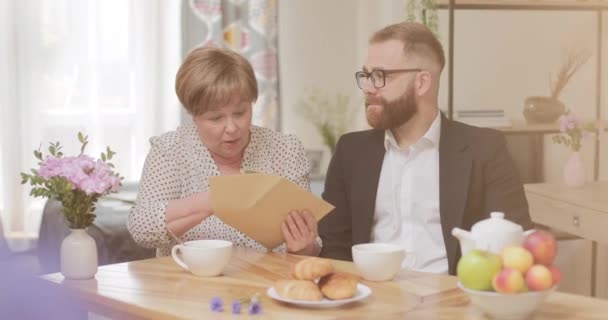 Homme d'affaires prospère donnant une grosse somme d'argent à sa mère retraitée surprise. Beau jeune homme aidant maman mature tout en apportant une enveloppe avec de l'argent. Concept de soins. — Video