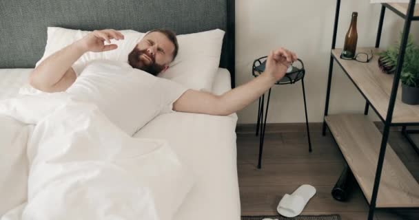 Fatigué jeune homme barbu se réveillant dans son lit et ayant la gueule de bois avec des maux de tête. Beau mec adulte dans les années 30 grimacant et touchant la tête avec les deux mains. Concept de mauvais matin. — Video