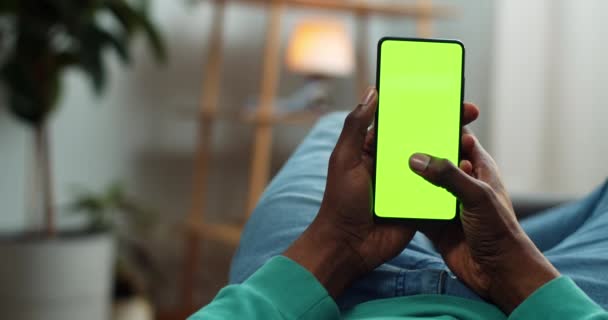 Cara em roupas casuais deitado no sofá e tocando smatphone simular exibição. Homem africano navegando rede enquanto olha para a tela verde. Conceito de chave croma. Aconchegante casa fundo . — Vídeo de Stock