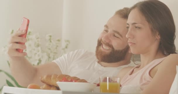 Close-up de casal sorrindo fazendo selfies e olhando para fotos enquanto deitado na cama de manhã cedo. Homem e mulher alegres usando se divertir durante o café da manhã no quarto. Conceito de lazer . — Vídeo de Stock