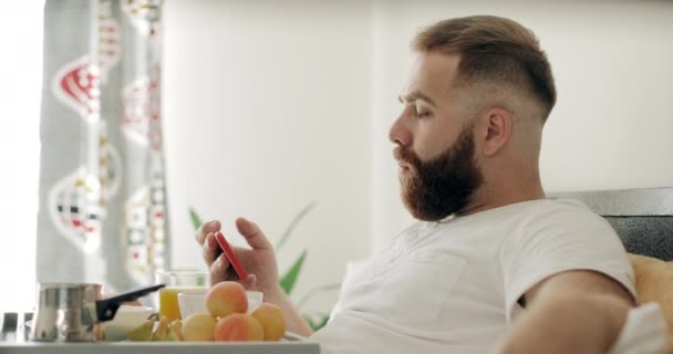 Młody brodaty mężczyzna żuje jedzenie oglądając filmy na swoim nowoczesnym smartfonie. Przystojny facet po trzydziestce patrzy na ekran telefonu podczas śniadania i siedzi z tacą z jedzeniem na łóżku. — Wideo stockowe
