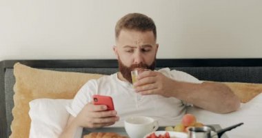 30 'lu yaşlarda sakallı bir adam akıllı telefon kullanıyor ve yatakta oturmuş meyve suyu içiyor. Bir adam tepsi dolusu yemekle otururken kahvaltı yapıyor. İnternet bağımlılığı kavramı.