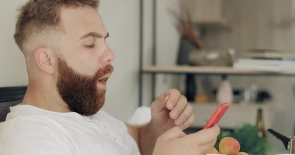 近视好看的男人在吃早餐的时候用智能手机打字的消息。留胡子的家伙一边一边一边一边咀嚼食物，一边用电话和坐着。技术与休闲的概念. — 图库视频影像