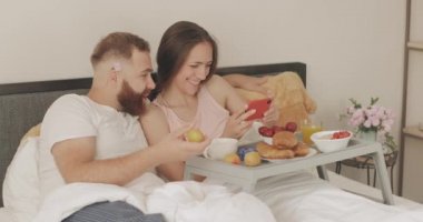 Neşeli genç aile telefon ekranına bakarken konuşuyor ve gülümsüyor. Kadın yatakta kahvaltıda akıllı telefon tutarken erkek meyveyle oynuyor. Boş zaman kavramı..