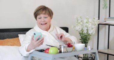 Görüntülü aramayı bitirip gülümseyen güzel, yaşlı bir kadın el sallıyor. Rob 'daki yaşlı kadın kahvaltı sırasında bacağında tepsiyle oturmuş video konferansı yapıyor..