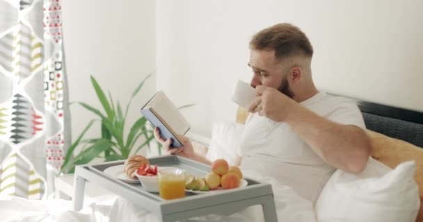 Vista lateral del chico concentrado en la mitad de los años 30 leyendo mientras desayunaba. Hombre barbudo guapo bebiendo café y pasando página de libro mientras está sentado en la cama. Concepto de ocio . — Vídeo de stock