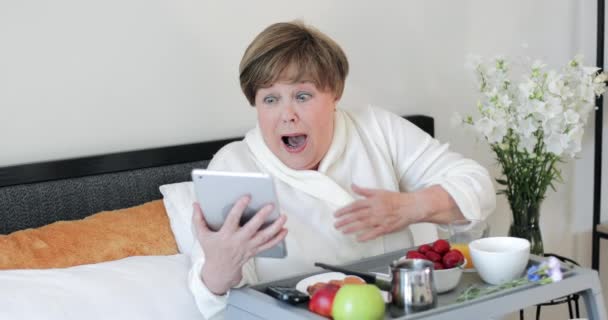 La anciana en Rob tiene malas noticias mientras mira la pantalla de gadget moderno. Mujer mayor sentada y desayunando en la cama mientras usa su tableta y se ve asustada. Concepto de emociones . — Vídeo de stock
