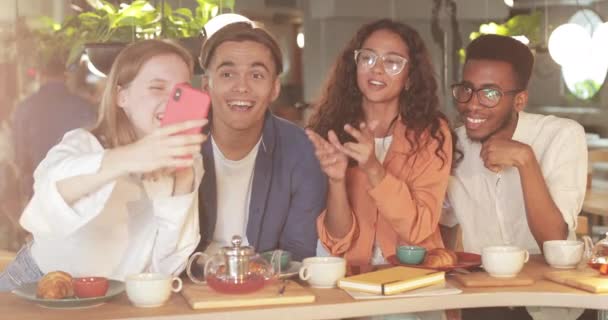 Joyful people making video message while laughing, waving and gesturing during lunch in cosy restaurant. Pretty woman holding mobilephone horizontally while looking at screen with friends. — Stock Video