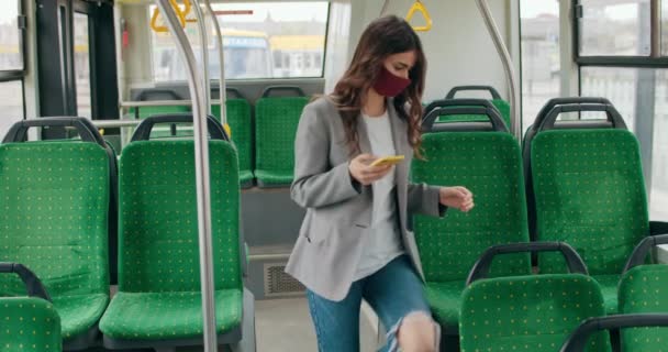 Brunetka dziewczyna w ręcznie robionej masce twarzy wchodzącej do autobusu i trzymając smartfona. Tysiącletnia kobieta siedzi na pustym siedzeniu podczas używania i przewijania ekranu telefonu. Pojęcie wirusa i pandemii. — Wideo stockowe