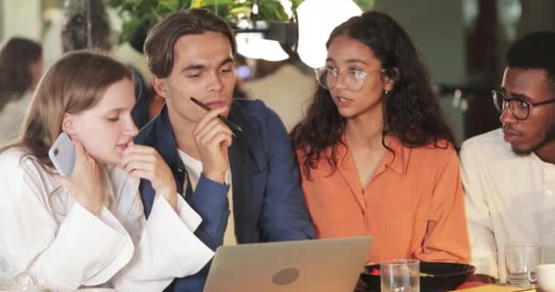 Jóvenes compañeros de trabajo creativos hablando y mirando la pantalla del ordenador portátil mientras están sentados en la cafetería. Diversos jóvenes concentrados discuten el proyecto. Acércate. Concepto de trabajo en equipo. En interiores . — Vídeos de Stock