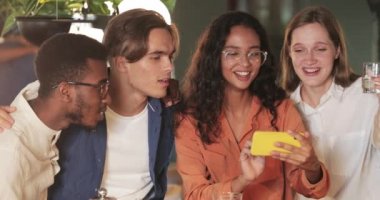 Neşeli bir milenyum kadını elinde akıllı telefonuyla arkadaşlarına komik fotoğraflar gösterip konuşuyor. Bir grup iş arkadaşı kahkahalarla gülüyor ve kafede takılırken telefon ekranlarına bakıyor..