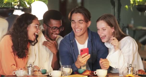 Jovem caucasiano mostrando seus amigos foto engraçada enquanto eles estão sentados no café acolhedor. Amigos alegres rindo e olhando para a tela do smartphone enquanto passam tempo juntos . — Vídeo de Stock