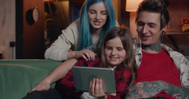 Сучасна сім'я сміється і торкається екрану, використовуючи планшет вдома. Міленіалістична мама, тато і їх дочка добре проводять час разом, сидячи на дивані з гаджетом. Концепція дозвілля . — стокове відео