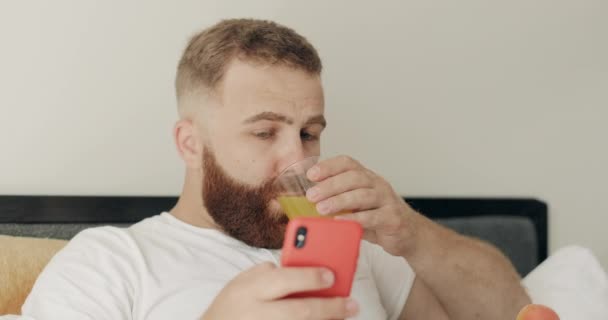 Närbild av skäggig ung man kvävs av juice samtidigt läsa chockerande nyheter på smartphone. Killen i 30-årsåldern gör förvånad ansikte medan du tittar på telefonskärmen och äter frukost i sängen. — Stockvideo