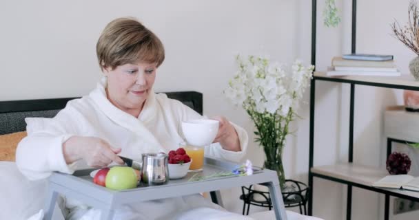 Een oude dame die koffie in de beker giet en drinkt terwijl ze met trey op haar benen zit. Vrolijke volwassen vrouw rob genietend goedemorgen tijdens het ontbijt op bed . — Stockvideo