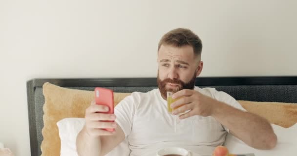 Hombre guapo bebiendo jugo y mostrando su desayuno mientras hace una videollamada. Chico feliz en los años 30 sonriendo y usando la cámara frontal del teléfono inteligente para la comunicación mientras está sentado en la cama . — Vídeos de Stock