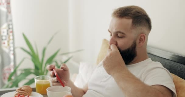 Pohled na pohledného muže, jak používá smartphone a jí, když sedí na posteli. Chlápek ve 30s se snídaní při posezení s tácem plným jídla a při pohledu na vertikální displej telefonu. — Stock video