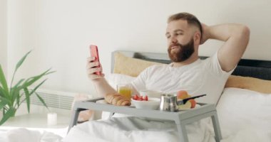Yakışıklı sakallı adam yatakta kahvaltı ederken video sohbeti yapıyor. Gülümseyen adam dizlerinde yemek tepsisiyle otururken akıllı telefonun ön kamerasını kullanıyor ve konuşuyor..