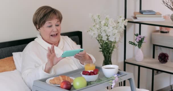 Madura mujer guapa haciendo foto de su desayuno mientras está sentado en la cama. Abuela de edad avanzada moderna utilizando el teléfono inteligente para hacer fotos y enviar mientras desayuna . — Vídeos de Stock