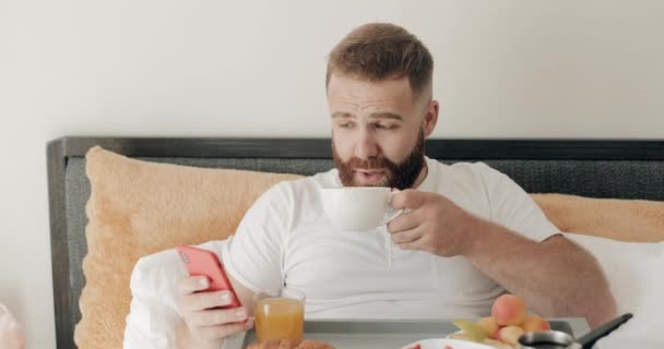 Vista frontal del joven barbudo usando su teléfono inteligente y sonriendo mientras toma café por la mañana. Chico guapo recibiendo mensaje divertido mientras desayuna y se sienta en la cama . — Vídeos de Stock