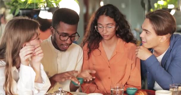 在现代咖啡馆里，一个戴着眼镜的非洲裔美国年轻人一边举手投足，一边向朋友们讲述有趣故事的作物景观。快乐的年轻人一边听一边笑。交流的概念. — 图库视频影像