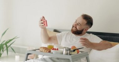 Mutlu sakallı adam, bacaklarında yemek tepsisiyle yatakta otururken selfie çekiyor. Gülümseyen adam sabah kahvaltı ederken akıllı telefonuyla poz veriyor ve fotoğraf çekiyor..