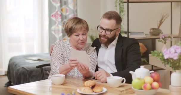 Goed uitziende volwassen moeder en volwassen zoon zitten en kijken naar oude foto 's tijdens het drinken van thee. Oude vrouw en jonge man herinneren zich oude tijden terwijl ze herinneringen deelden. Gezellige thuis achtergrond. — Stockvideo