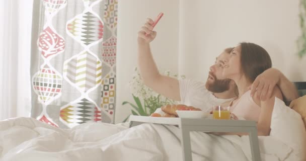 Couple souriant amoureux faire selfies tout en étant couché sur le lit tôt le matin. Homme et femme joyeux en utilisant la caméra frontale du smartphone tout en prenant le petit déjeuner dans la chambre. Concept de loisir. — Video