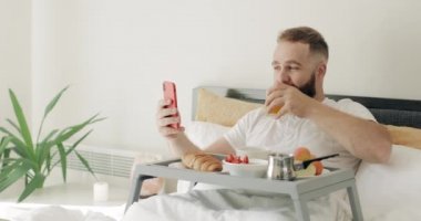 Mutlu sakallı adam meyve suyu bardağını alkışlıyor ve video sohbeti yaparken kahvaltı yapıyor. 30 'lu yaşlarda bir adam dizlerinin üzerinde yemek dolu tepsiyle otururken akıllı telefon kullanıyor..