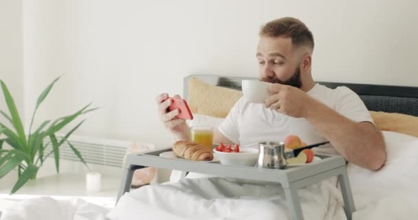 Bärtiger Mann, der Kaffee trinkt und lustige Videos anschaut, während er auf dem Bett liegt.Fröhlicher Typ in den 30er Jahren, der auf den Smartphone-Bildschirm schaut, ihn beim Frühstück senkrecht hält und lacht. — Stockvideo