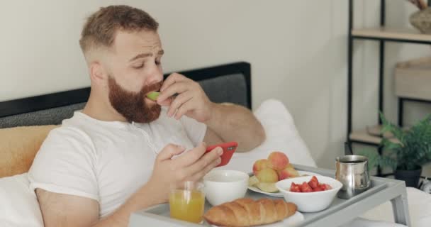 Красивий молодий чоловік має здоровий сніданок, використовуючи свій смартфон. Бородатий чоловік дивиться на екран телефону і читає новини, сидячи на ліжку з підносом, повним їжі . — стокове відео