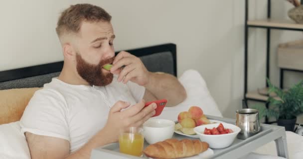 Handsome young man having healthy breakfast while using his smartphone. Bearded man looking at phone screen and reading news while sitting on bed with tray full of food. — Stock Video