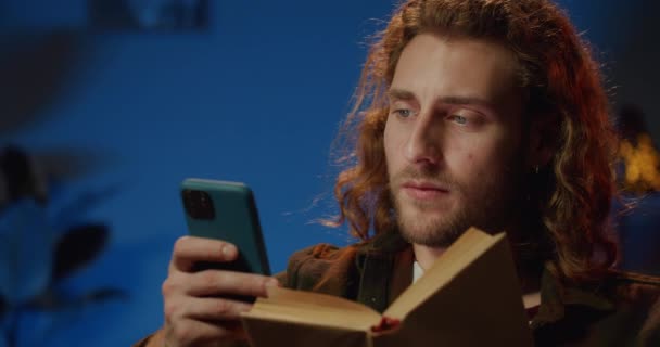 Close-up zicht op knappe jongeman met moderne smartphone in de ene hand en geopend boek in de andere. Millennial serieuze man met behulp van telefoon en literatuur te lezen. Achtergrondinformatie. — Stockvideo