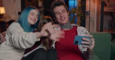 Dövmeli ve piercingli neşeli ebeveynler güzel kızlarıyla kanepede otururken görüntülü görüşme yapıyorlar. Modern aile evde iletişim kurmak için telefon kullanırken el sallıyor ve konuşuyor..