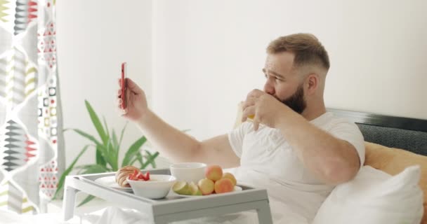 Vue latérale du gars joyeux ayant une conversation vidéo tout en étant assis et en prenant le petit déjeuner au lit. Homme parlant et souriant tout en utilisant la caméra frontale du smartphone. Concept de communication. — Video