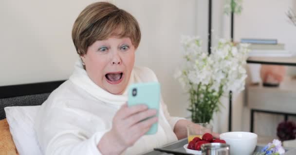 Närbild av äldre kvinna som har goda nyheter och kan inte tro sina ögon när du använder smartphone. Gammal pensionerad dam gör förvånad ansikte medan du äter frukost och tittar på telefonskärmen. — Stockvideo