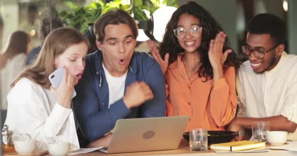 Des jeunes excités ayant de bonnes nouvelles et se réjouissant tout en étant assis dans un café. Jeunes souriants collègues divers regardant heureux tout en utilisant un ordinateur portable. Concept de travail d'équipe et de victoire. — Video