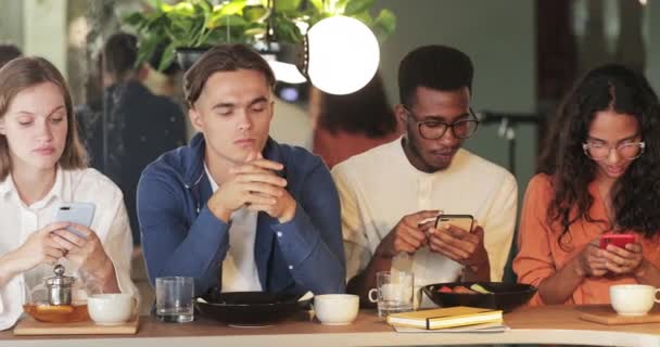 Amigos del milenio usando sus teléfonos mientras pasan tiempo juntos en la cafetería. Chico joven que parece aburrido mientras está sentado entre gadget adictos multiétnicos con teléfonos inteligentes . — Vídeo de stock