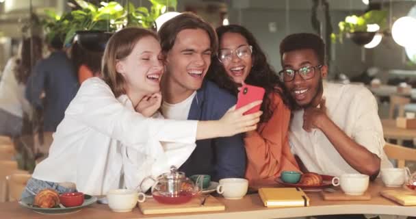 Un colega alegre mirando la pantalla del teléfono mientras pasa un buen rato durante el almuerzo. Jóvenes amigos felices viendo fotos en el teléfono inteligente mientras están sentados en la mesa en el restaurante moderno . — Vídeos de Stock