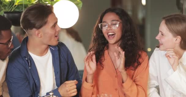 Hezká dívka v brýlích mluví, usmívá se a objímají své přátele v moderní kavárně. Oříznout pohled na mnohonárodnostní mladé lidi, jak si povídají a smějí se. Koncept komunikace. — Stock video
